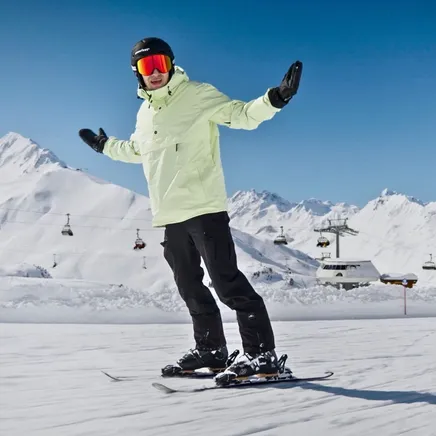 Un skieur en veste vert clair et lunettes orange, les bras écartés, sur une piste enneigée avec des montagnes et télésièges sous un ciel bleu.