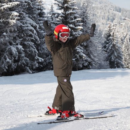 Un enfant skie sur une piste enneigée, vêtu d'un casque rouge et d'une combinaison marron, les bras levés pour l'équilibre. Des sapins en arrière-plan.