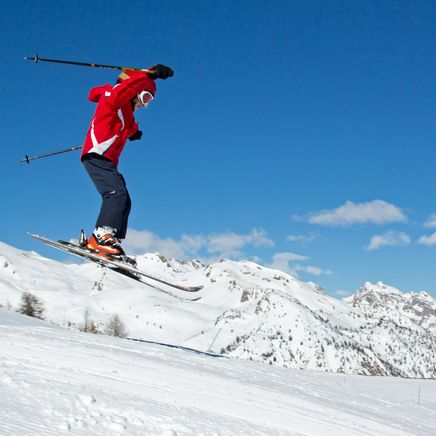 Skieur en veste rouge en plein saut au-dessus d'une piste de ski, avec des montagnes enneigées et un ciel bleu clair en arrière-plan.