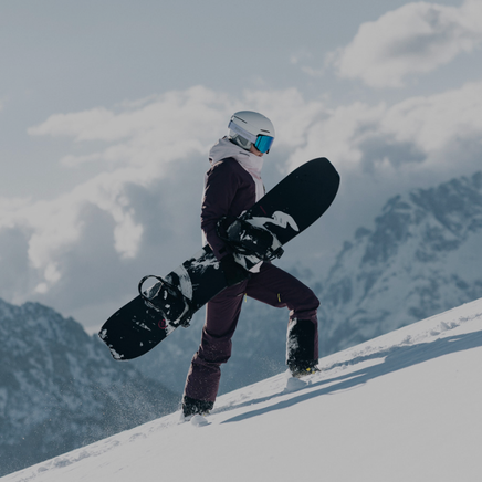 Une snowboardeuse en tenue d'hiver, casque et masque, monte une pente enneigée avec sa planche. Montagnes majestueuses.