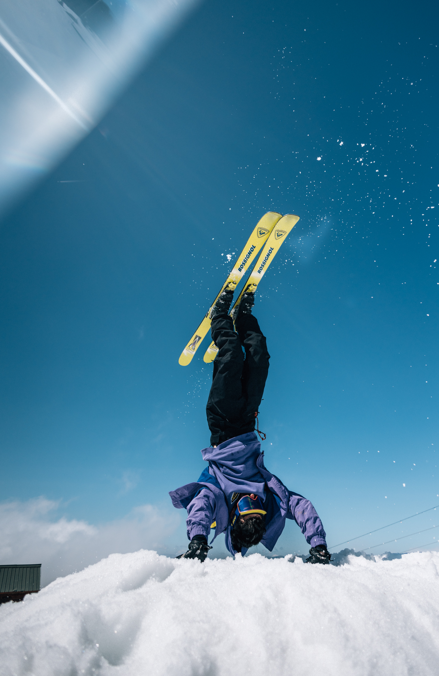 Skieur en équilibre sur les mains dans la neige, skis jaunes pointant vers le ciel bleu clair.