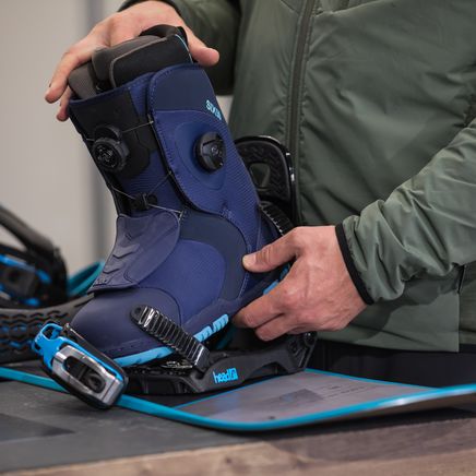 Une personne place une botte de snowboard bleue à laçage BOA dans une fixation sur une planche de snowboard turquoise.