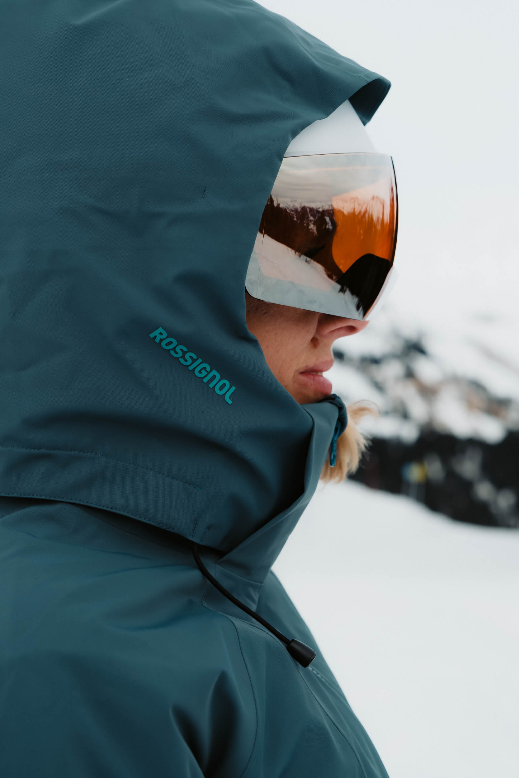 Profil d'une personne en veste de ski Rossignol bleu-vert et masque reflétant des montagnes enneigées.