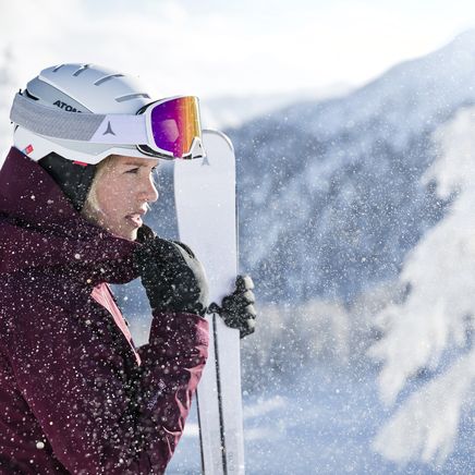 Femme skieuse en casque et lunettes, tenue bordeaux, tenant des skis sous une chute de neige, avec montagnes enneigées en arrière-plan.