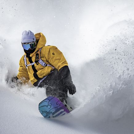 Un snowboarder en veste jaune et bonnet blanc fait un virage serré, soulevant de la neige poudreuse sur une pente enneigée.