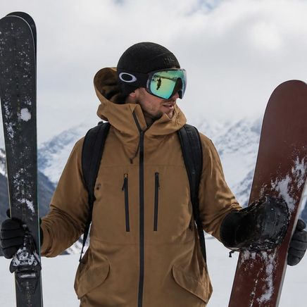 Skieur en tenue hivernale, avec masque et bonnet, tenant des skis et un snowboard devant des montagnes enneigées.