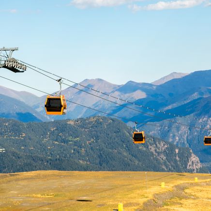 Des télécabines oranges traversent un paysage de montagne avec des pentes herbeuses dorées, des forêts et des sommets lointains.