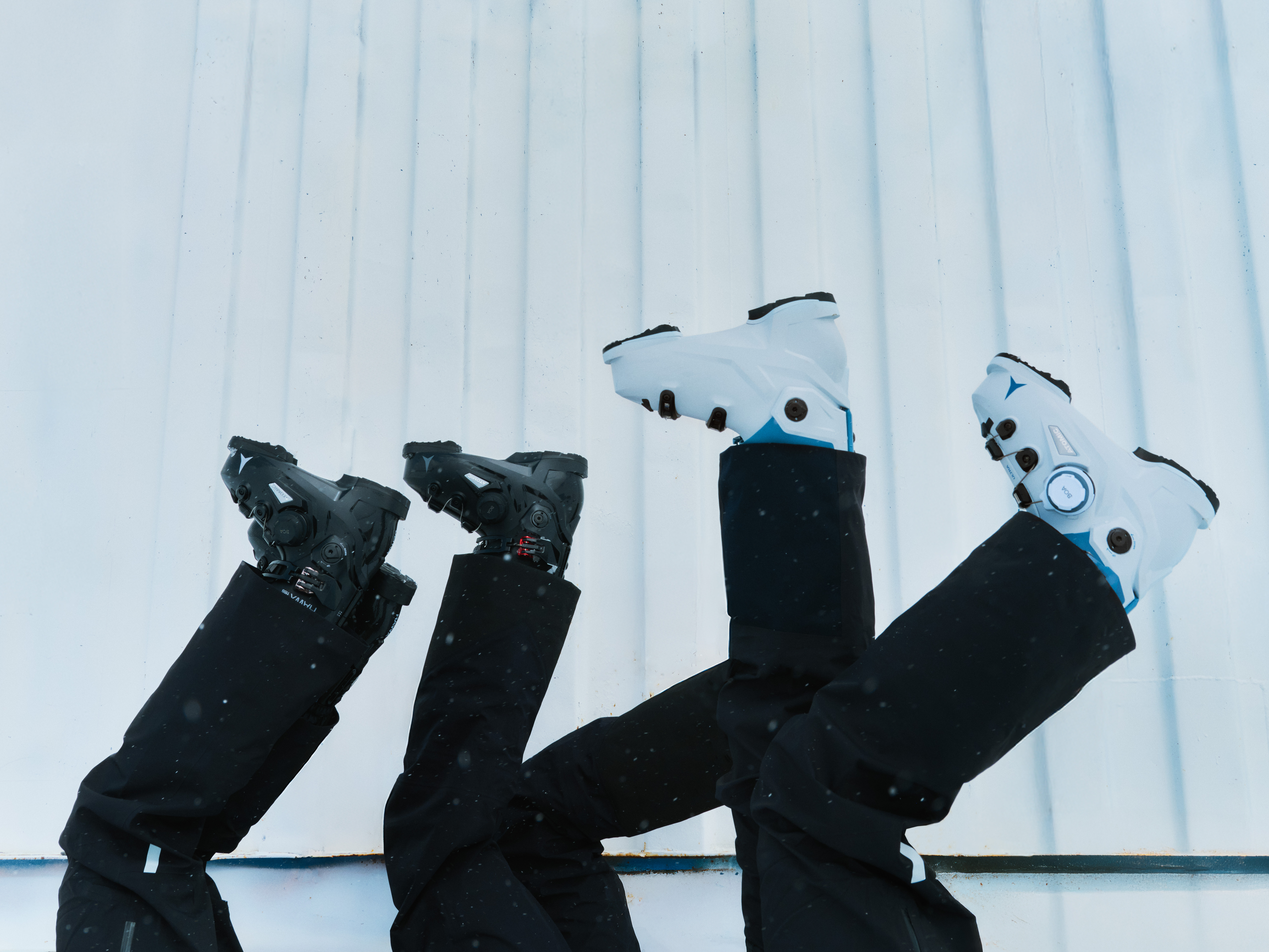Quatre paires de jambes en pantalons et bottes de ski (deux noires, deux blanches) levées contre un mur bleu pâle.