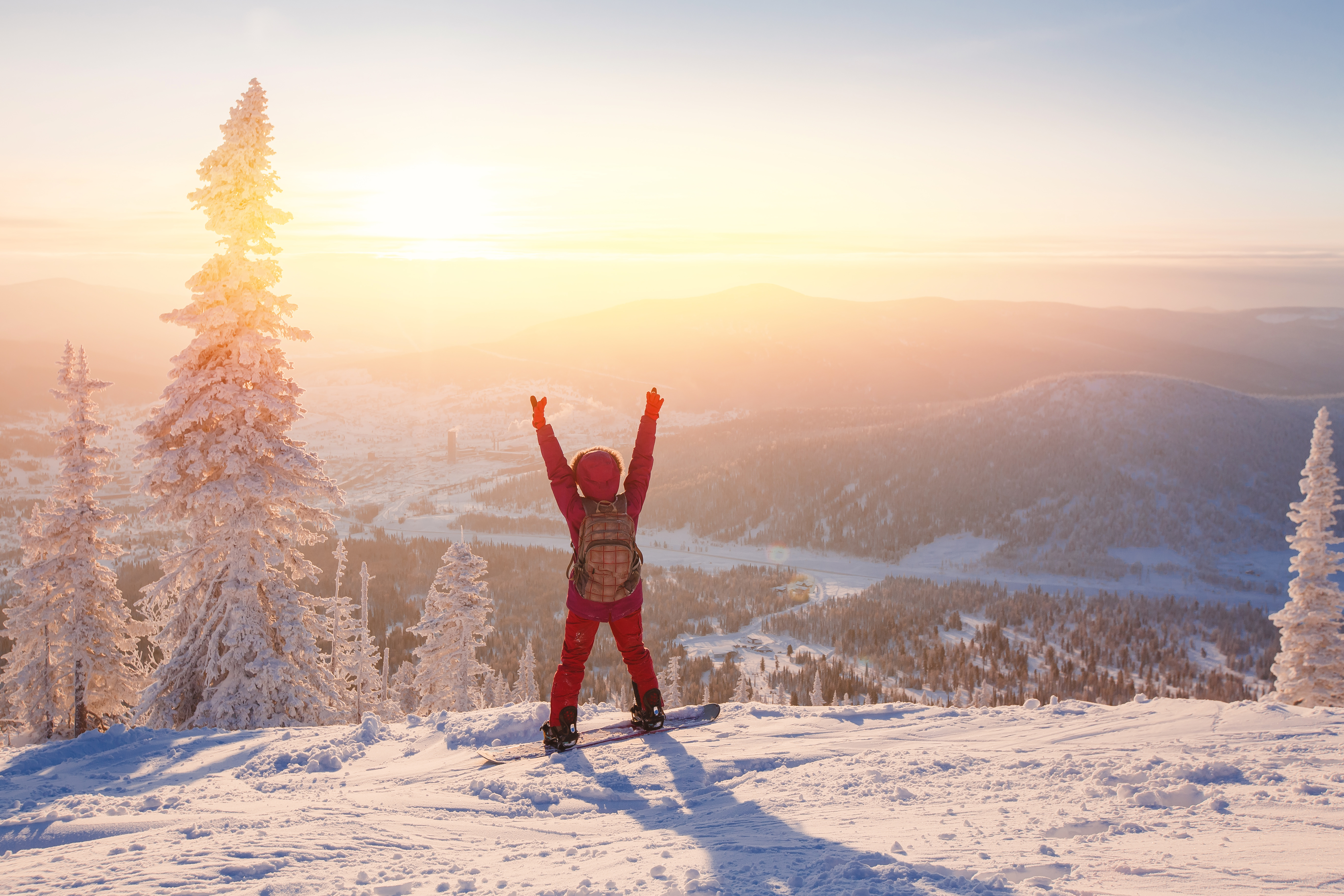 Snowboarder les bras levés sur un sommet enneigé au lever du soleil, avec une vaste vallée forestière et des sapins givrés en arrière-plan.