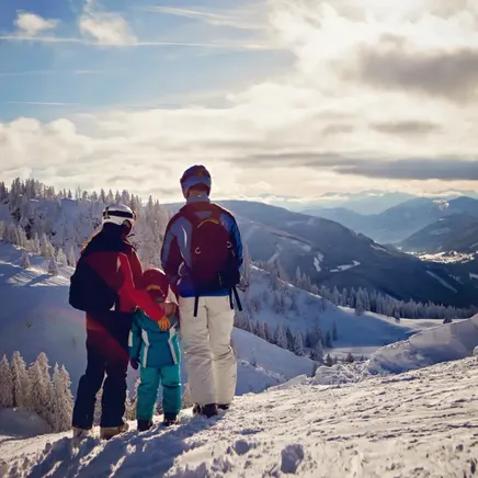 Une famille (2 adultes, 1 enfant) admire un vaste paysage hivernal de montagnes enneigées et de vallées sous un ciel lumineux.