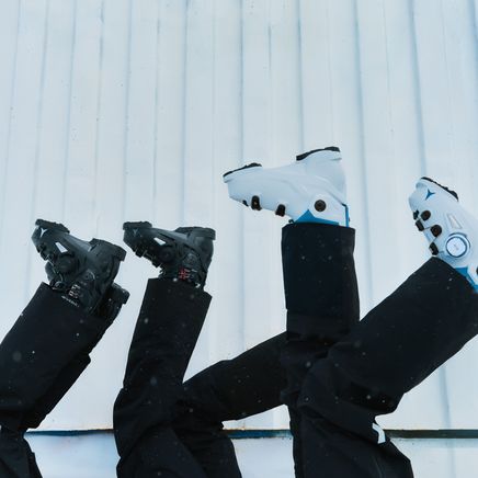 Quatre jambes en pantalons de ski noirs, chaussées de bottes (deux noires, deux blanches), pointent vers le haut devant un mur blanc.