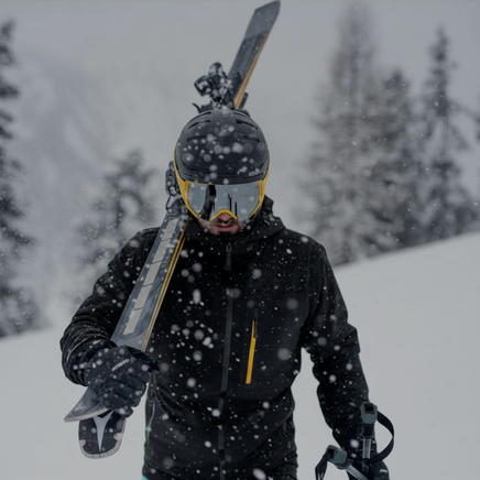 Un skieur équipé porte ses skis sur l'épaule sous une neige abondante en montagne.