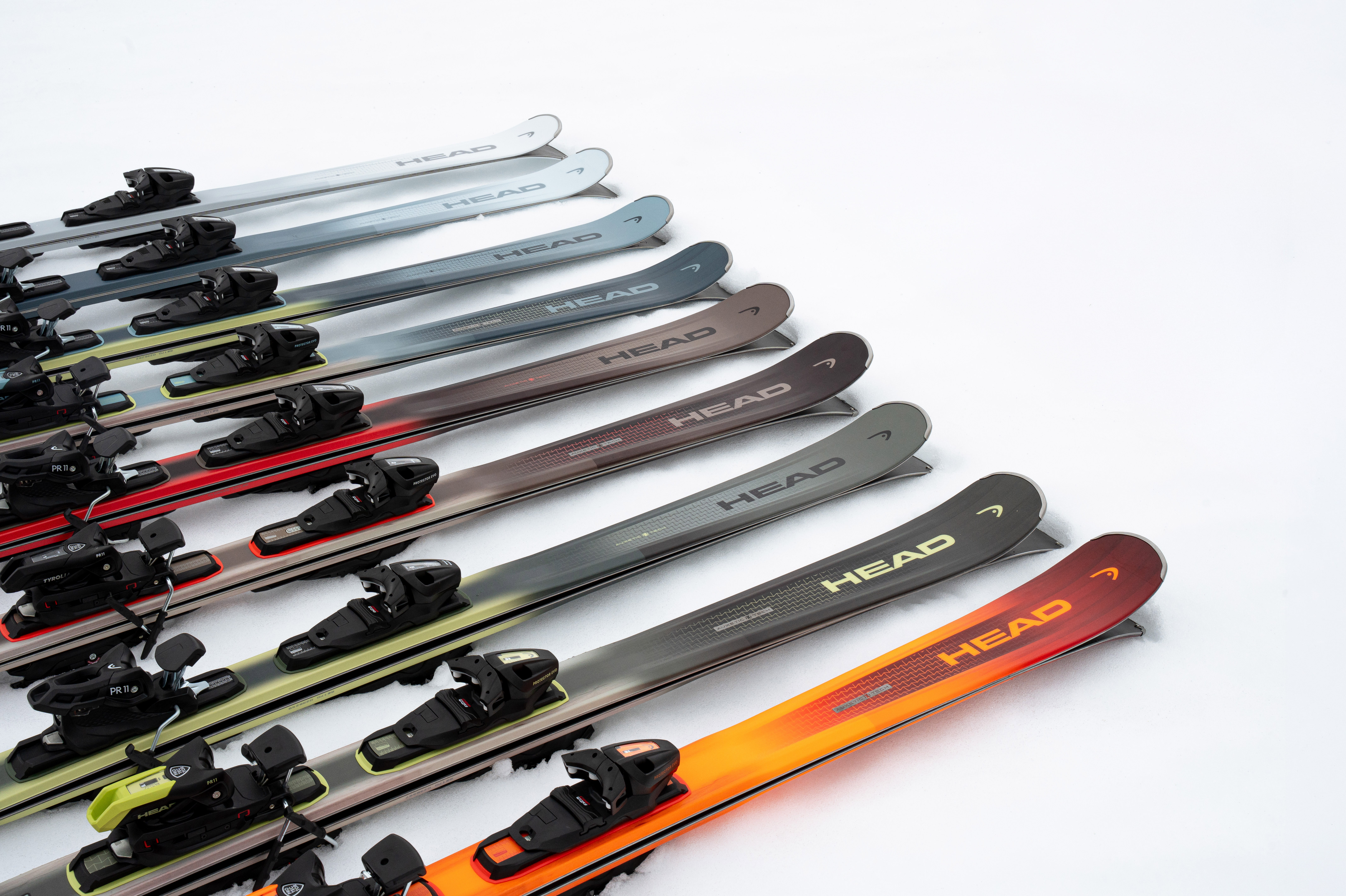Plusieurs paires de skis Head de couleurs variées avec fixations, alignées sur la neige, créant un dégradé de teintes.