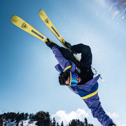 Skieur en veste violette et jaune réalisant un saut acrobatique spectaculaire, skis jaunes en l'air, sous un ciel bleu et soleil éclatant en montagne.