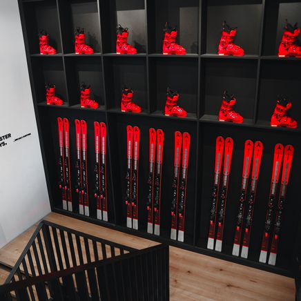 Bottes et skis de ski rouges alignés sur des étagères noires, avec la citation "ALWAYS BE FASTER THAN THE BOYS" sur un mur blanc à côté d'un escalier.
