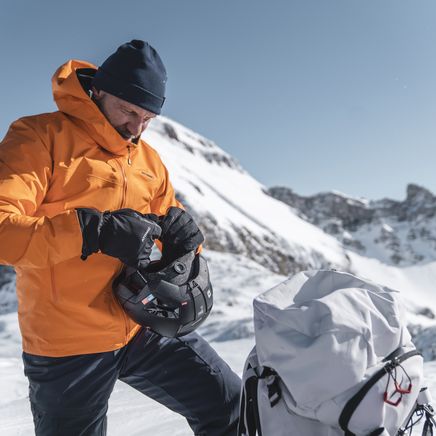 Skieur en veste orange et bonnet, ajustant son casque devant des montagnes enneigées. Un sac à dos blanc est à côté de lui.