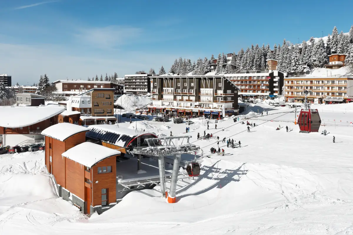Station de ski ensoleillée avec immeubles enneigés, télécabines rouges, nombreux skieurs sur les pistes et forêt de sapins.