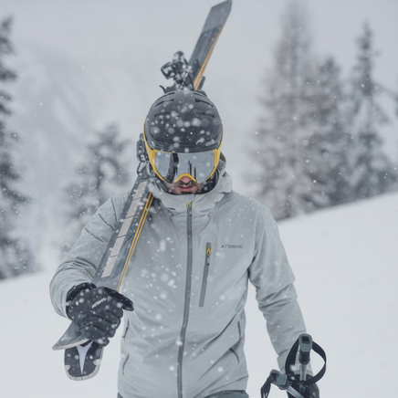 Skieur en tenue grise et jaune, skis sur l'épaule, bâtons en main, marchant dans un paysage montagneux enneigé sous des flocons.
