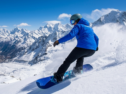 Un snowboarder en veste bleue, casque et lunettes dévale une pente enneigée. De la neige jaillit. Montagnes blanches et ciel bleu.