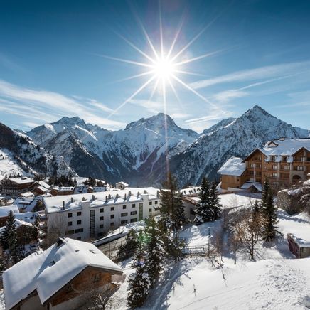 Village de montagne enneigé sous un soleil éclatant, avec des chalets et des sommets majestueux. Ciel bleu limpide.