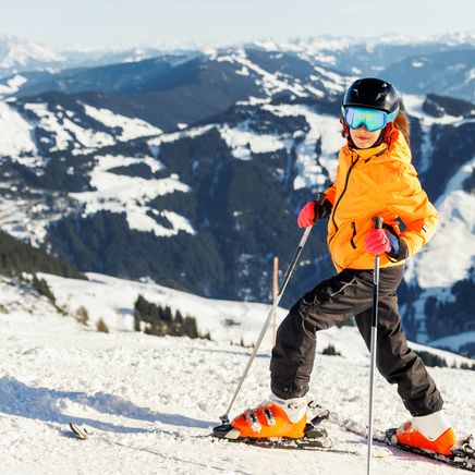 Enfant skieur en veste orange, casque et lunettes bleues sur une piste enneigée avec des montagnes en arrière-plan sous un ciel ensoleillé.