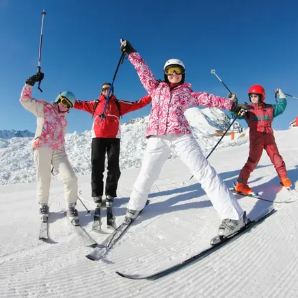 Quatre personnes en tenue de ski sourient et posent avec leurs bâtons levés sur une piste enneigée, sous un ciel bleu.