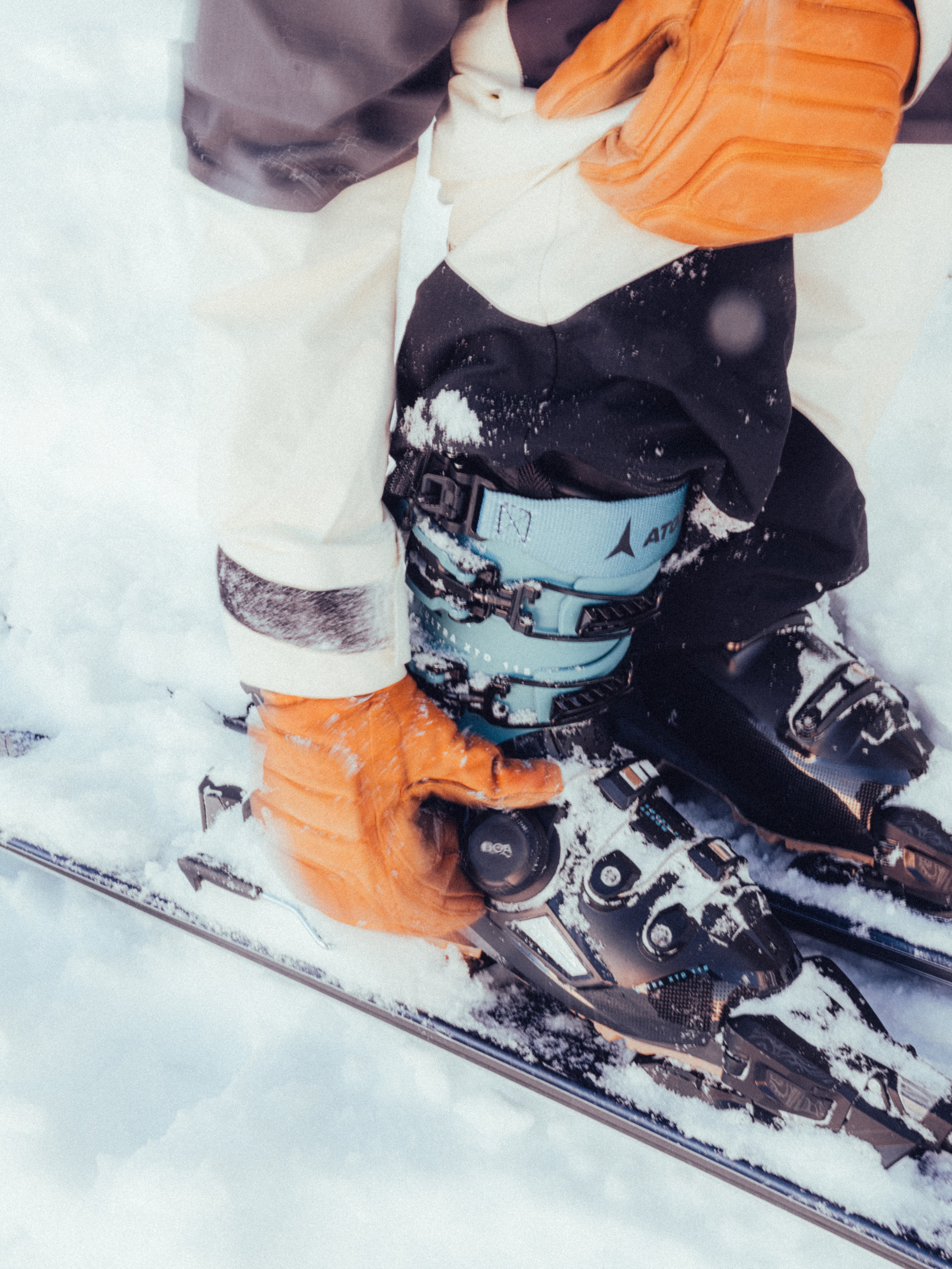 Une personne gantée d'orange ajuste sa chaussure de ski Atomic bleue et noire, couverte de neige fraîche.