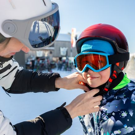 Un adulte ajuste le casque d'un enfant souriant en tenue de ski sur une piste enneigée.