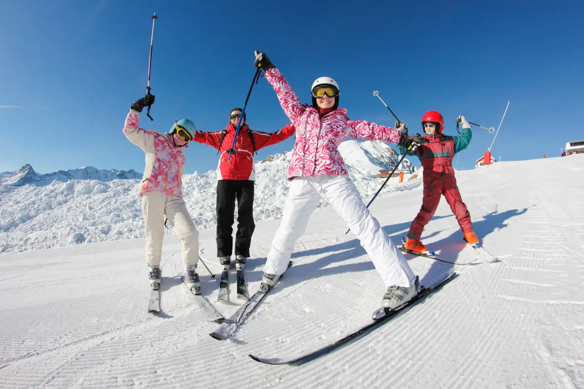 Quatre skieurs joyeux posent sur une piste enneigée, ciel bleu et montagnes en arrière-plan.