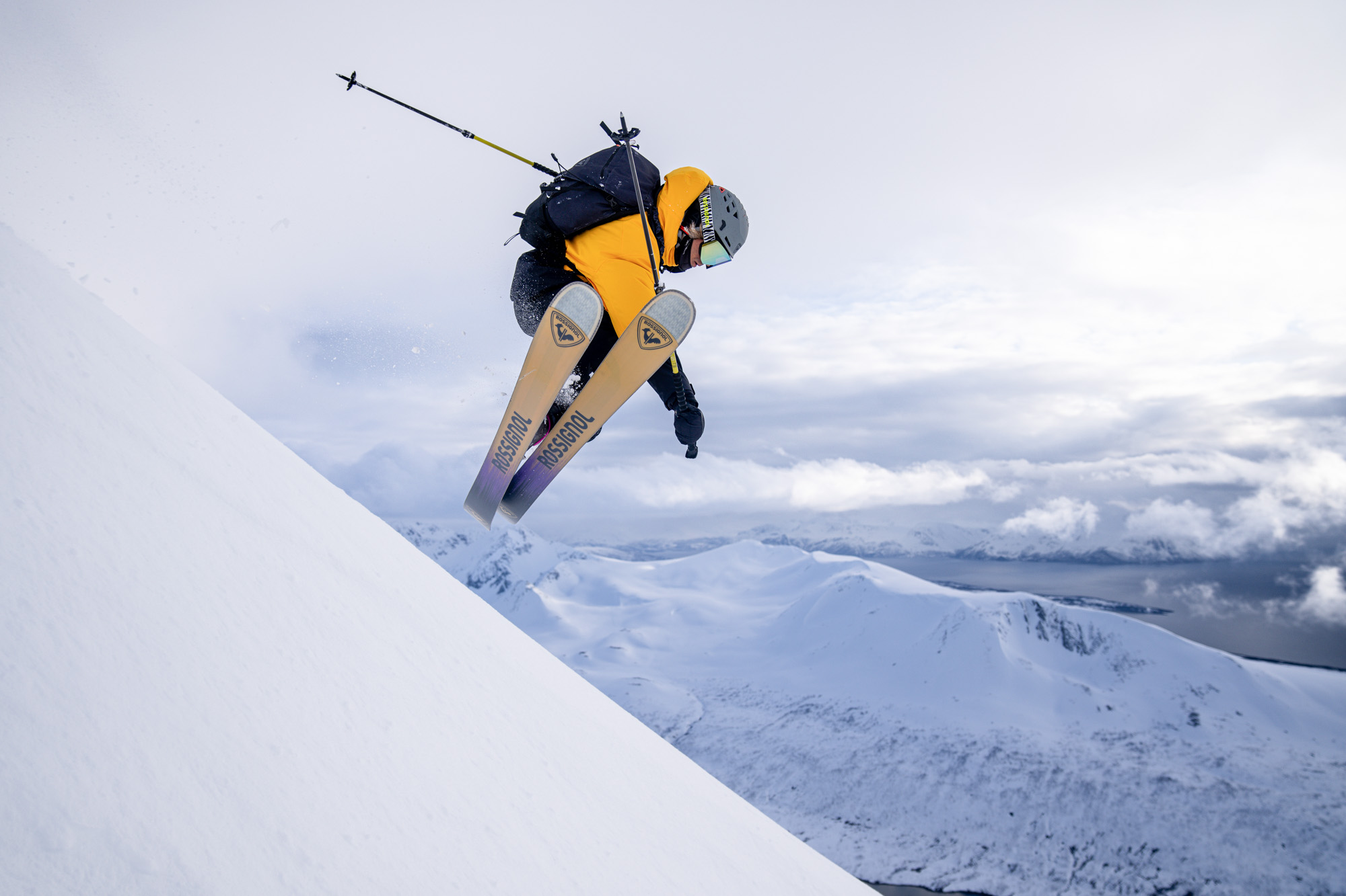 Skieur en veste jaune et sac à dos noir sautant sur une pente enneigée, montagnes et fjord sous un ciel nuageux.